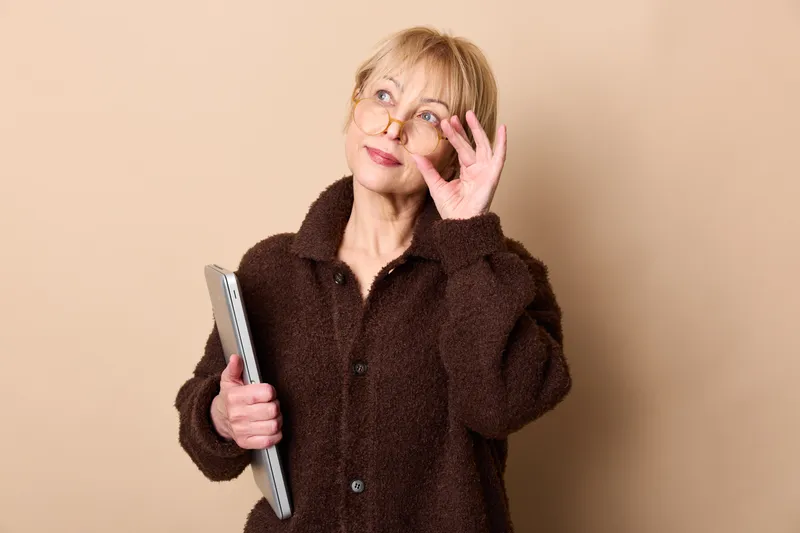 Woman holding smartphone, smiling in portrait, wearing brown fuzzy coat, posing against beige background, hand forming a peace sign, calm self confidence, soft studio lighting, everyday communication