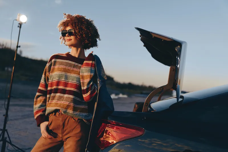 Woman with curly hair stands beside open car trunk at sunset, outdoor lifestyle and travel mood, warm evening light, casual sweater and sunglasses, travel preparation, cinematic background