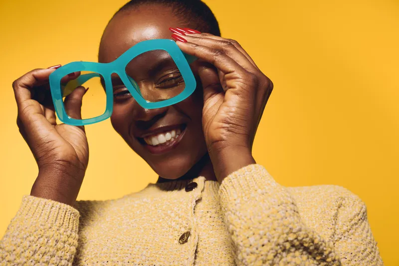 Happy African woman with dark skin smiles joyfully while holding oversized blue glasses in front of her face. Yellow background and cozy beige sweater create a warm, playful mood.