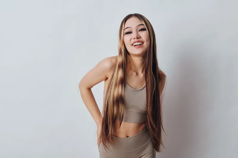 Young woman with long braided hair wearing beige sportswear smiling and posing with hands on hips against light gray background in studio portrait.