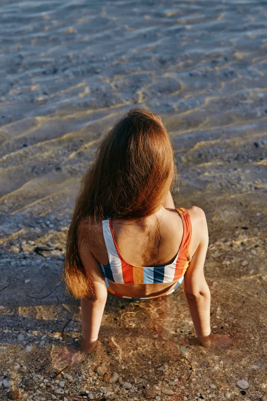 A girl sits in shallow water at the beach, back turned to the camera, wearing a striped swimsuit. The sunlit scene captures calm, carefree coastal moments and a tranquil seaside mood.
