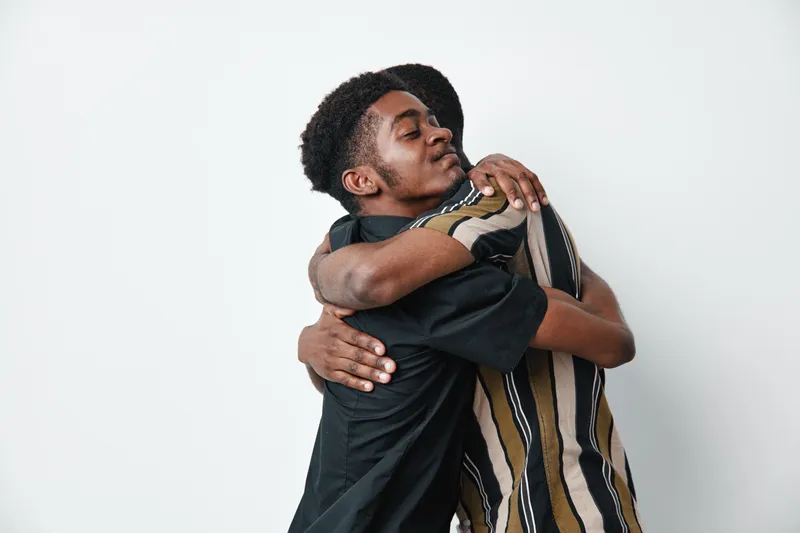 Two men with dark skin hugging each other in a friendly embrace on white background. African ethnicity, casual clothing, emotional connection, support and friendship concept.