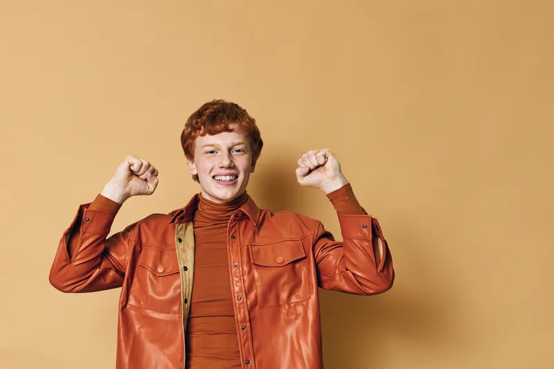Happy young man with red hair wearing brown leather jacket and turtleneck smiling and raising fists in celebration against beige background in studio portrait.