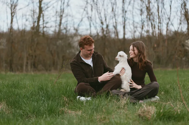 Casual outdoor lifestyle with a cheerful couple sitting in a field, playing with a goat kid, warm afternoon light, relaxed smiles, gentle interaction, natural setting, candid moment of joy and