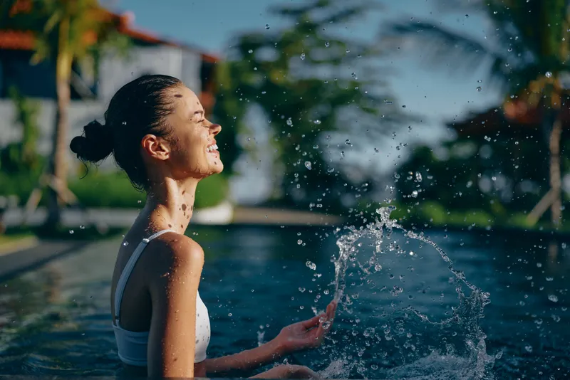 A carefree woman enjoys a splash by the pool, smiling as water sprays in a bright outdoor setting. A lifestyle moment of relaxation, warmth, and refreshing leisure by water.