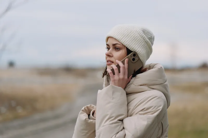 Woman in winter coat talks on a phone outdoors, wearing a beanie, standing beside a rural road in overcast weather.