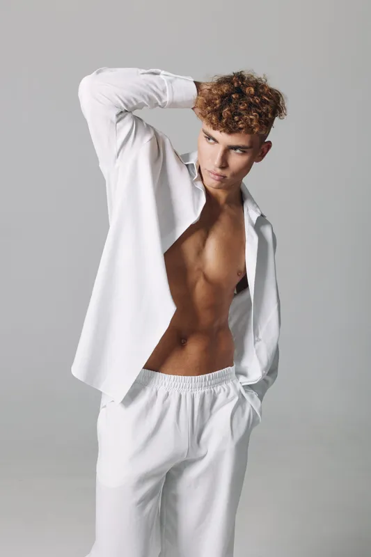 Young man with curly hair posing in white open shirt and pants against gray background, showing toned body and casual fashion style in studio lighting.