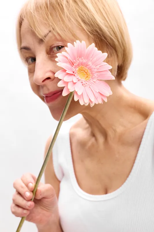 Blonde woman portrait holding pink gerbera flower near face, soft closeup beauty, gentle smile, white background, natural skin, spring freshness, minimal calm studio