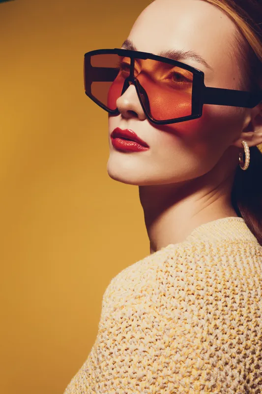 Stylish woman with oversized sunglasses and red lipstick poses against yellow background. Fashion portrait with trendy accessories and warm lighting for beauty and style concept.