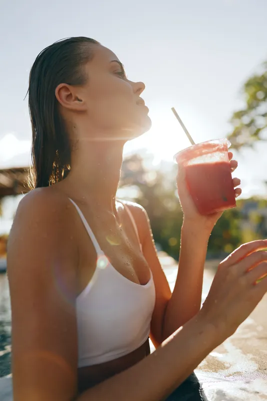 Relaxed woman by the pool enjoys a refreshing red drink in sunlight, wearing a white swimsuit, capturing a carefree summer moment outdoors with breeze and glow elevating mood.