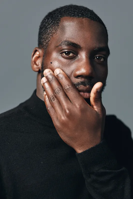 African man with dark skin and short hair in creative conceptual visual style wearing black sweater touching face with hand against gray background. Portrait with thoughtful expression and detail.
