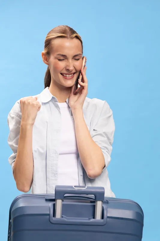 Happy woman with suitcase talks on smartphone, celebrating travel plans. Casual outfit and light blue background create positive mood of vacation, journey, and communication.