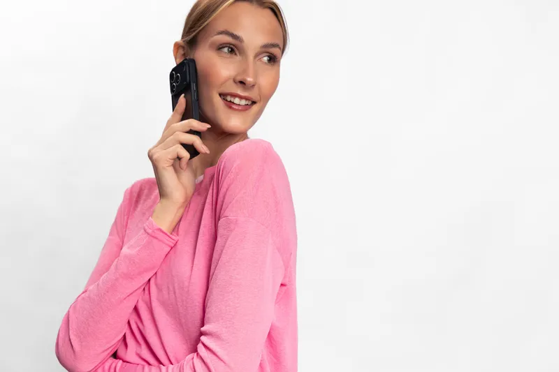 Young woman in pink sweater holds phone near ear and smiles looking away. Isolated on white background. Concept of communication, happiness, and casual conversation.