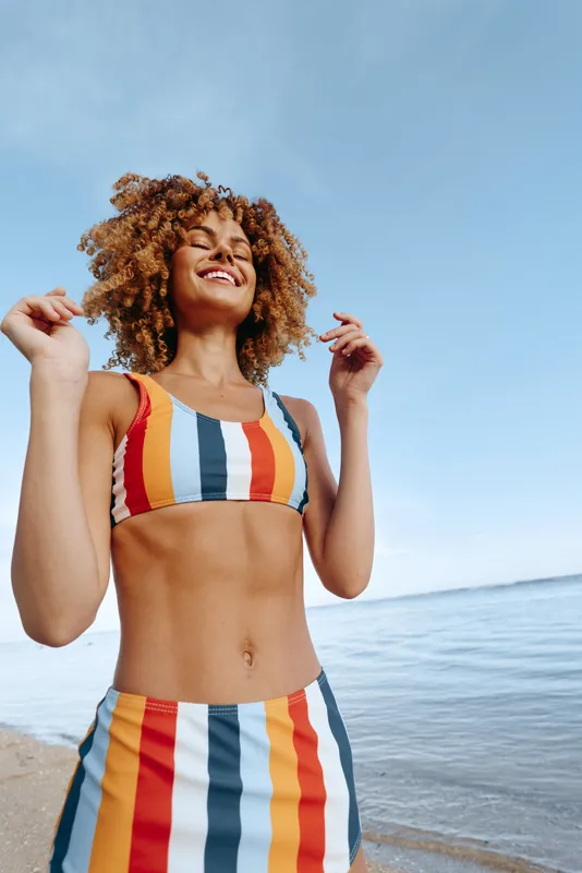 Woman with curly hair in striped swimsuit at the ocean beach on a sunny summer day, smiling and adjusting her hair, relaxed vacation mood, wellness concept