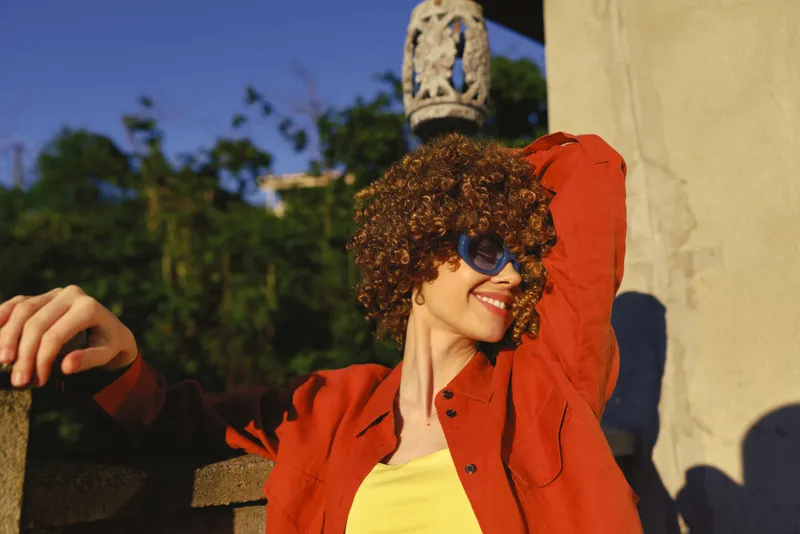 Cheerful woman outdoors, relaxed lifestyle moment, wearing orange jacket and yellow top, sunglasses on, sunlit breeze, joyful smile, lush greenery background, vibrant mood