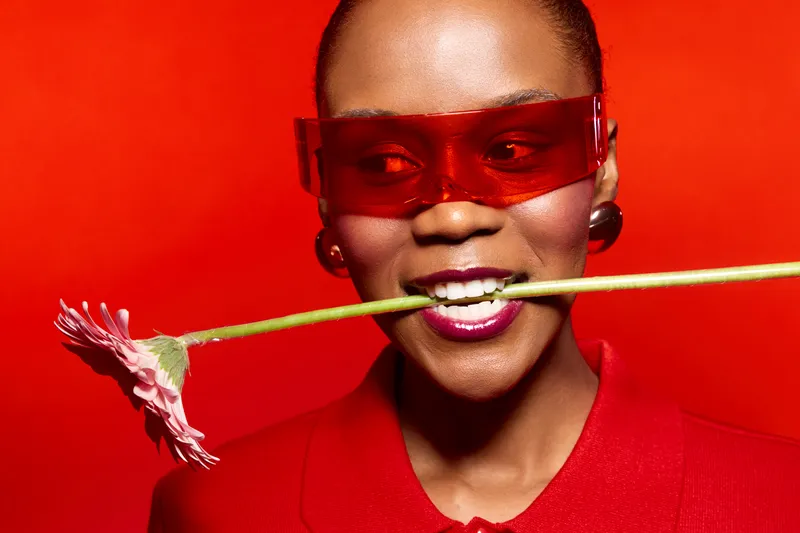 Portrait of smiling black woman in red sunglasses and blazer holding pink flower in teeth on red background. Concept of fashion, style, beauty, and confident African ethnicity model.