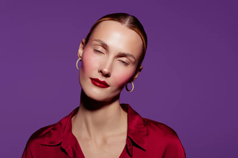 Portrait of a woman with closed eyes wearing red lipstick and earrings against purple background. Concept of beauty, fashion, makeup, and elegance in studio lighting.
