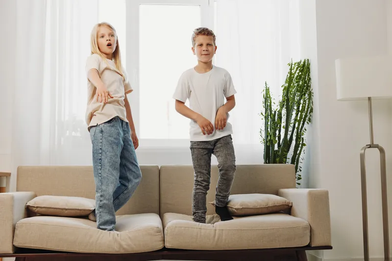 Two children stand on a beige sofa in a bright living room with white curtains and green plants. The girl points forward while the boy looks ahead calmly.