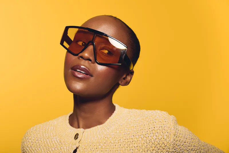 Portrait of a confident black woman with dark skin wearing oversized sunglasses and a beige knitted sweater. Yellow background enhances the vibrant and stylish concept of fashion and beauty.