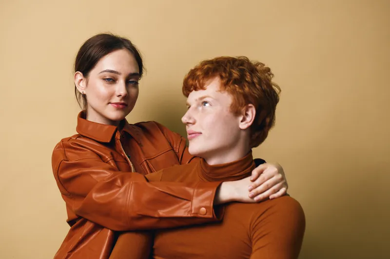 Young couple embracing and looking at each other in cozy autumn outfits, woman in leather jacket and man in turtleneck against beige background, warm and intimate moment.