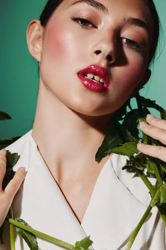 Close-up of a woman with vibrant makeup wearing white clothing holding green plant stems. Glossy lips and soft pink blush create a bold beauty fashion portrait.