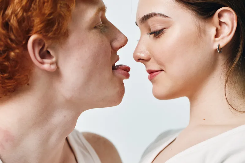Young couple closeup with playful expression, man sticking tongue out near woman face with smile, light background, intimacy and affection moment.