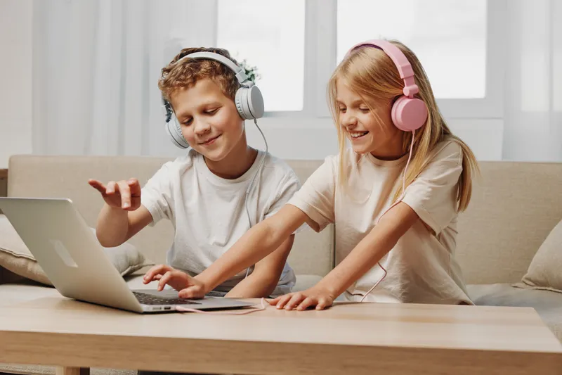 Two children wearing headphones enjoy music and games on a laptop, sitting on a sofa in a bright room with casual clothing and smiles.