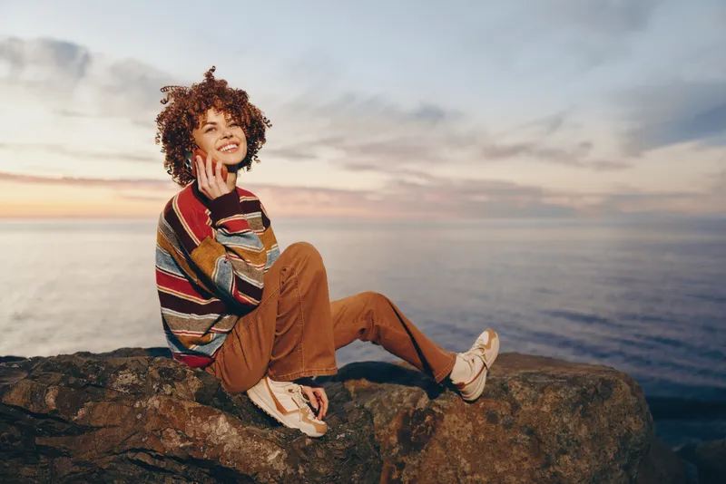 Woman lifestyle portrait, seated on a rocky cliff above the ocean during sunset, curly hair, casual clothing, relaxed smile, leisure mood, calm nature, waves and open sky