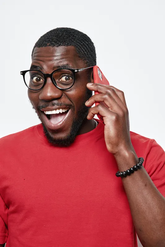 African man with dark skin wearing glasses and red sweater talking on smartphone with excited expression isolated on white background. Communication, technology and happiness concept.
