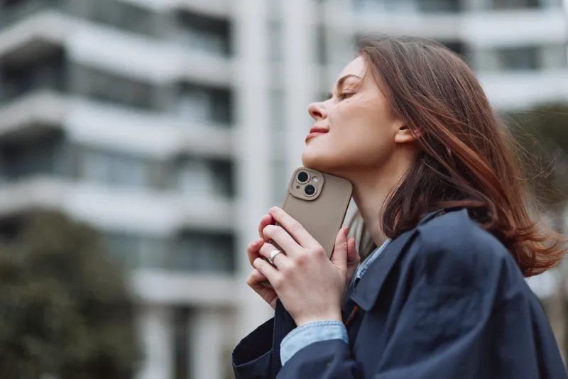 Young woman with brunette hair enjoys a peaceful moment outdoors while holding a smartphone close to her face with closed eyes and a slight smile.