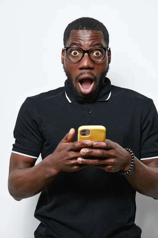 African American man with dark skin and glasses looking surprised while holding yellow smartphone in black polo shirt on white background. Shocked expression and casual style.