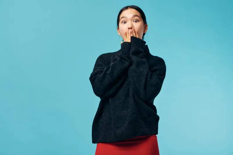 Woman wearing black sweater and red skirt standing in front of blue background with hands covering mouth expressing surprise or shock. Portrait of emotional young female with casual outfit.