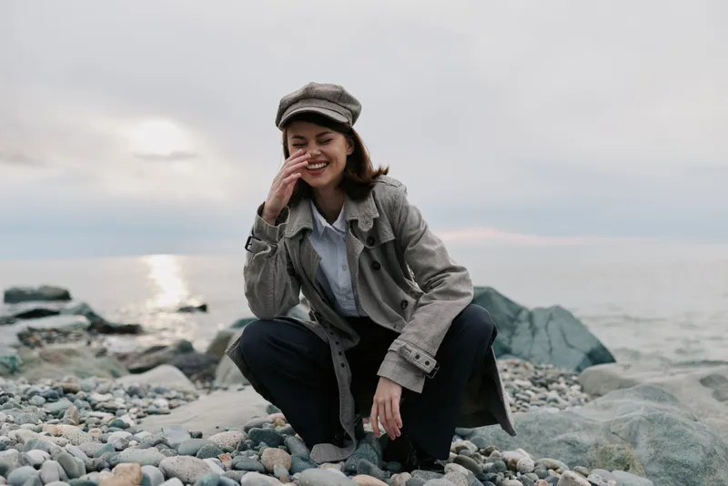 Young woman wearing a hat and coat squats on a rocky beach near the sea, smiling and covering her face with one hand. Overcast sky with soft natural light creates a calm mood.