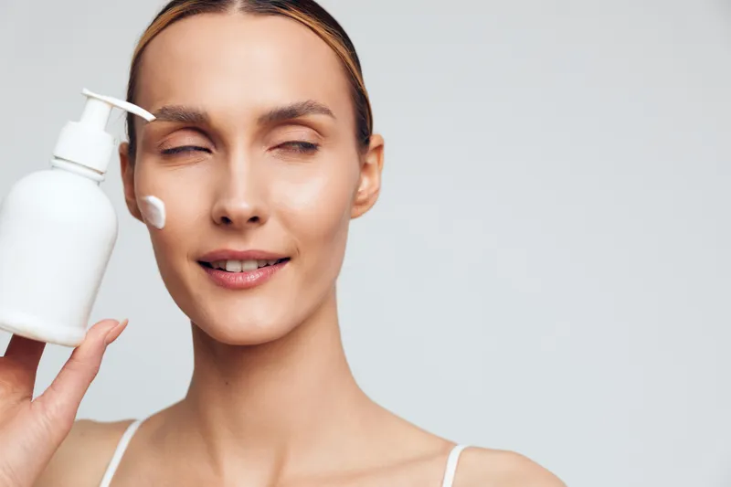 Young woman with clear skin holding white lotion bottle applying cream on cheek, smiling with closed eyes. Concept of skincare, beauty routine, hydration, and self-care on light background.