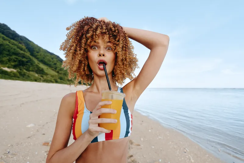 Woman with curly hair drinks juice on the beach by the ocean in summer, hydration and wellness lifestyle, outdoor sunlit beach scene, playful expression, holding straw bottle, ocean breeze