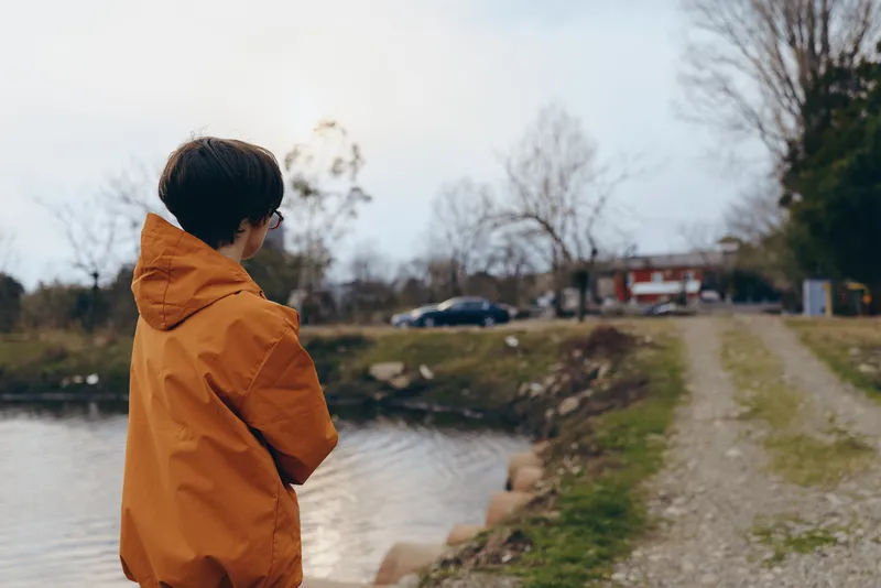 Woman outdoor lifestyle, orange hooded coat, standing by river bank, side view, park path, leafless trees, cloudy daylight, walking calm nature mood, quiet fresh air, countryside view