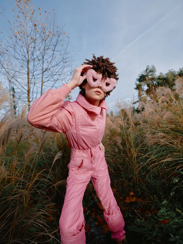 Child in a pink jumpsuit wearing a feathered mask stands in tall grass outdoors, playful pose under clear sky, autumn scenery, creative costume and nature setting