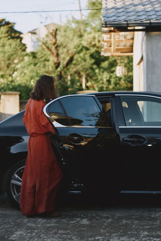 Candid woman in orange dress opening a car door outdoors, sunlit driveway scene, relaxed lifestyle moment, warm tones, contemporary fashion portrait natural light