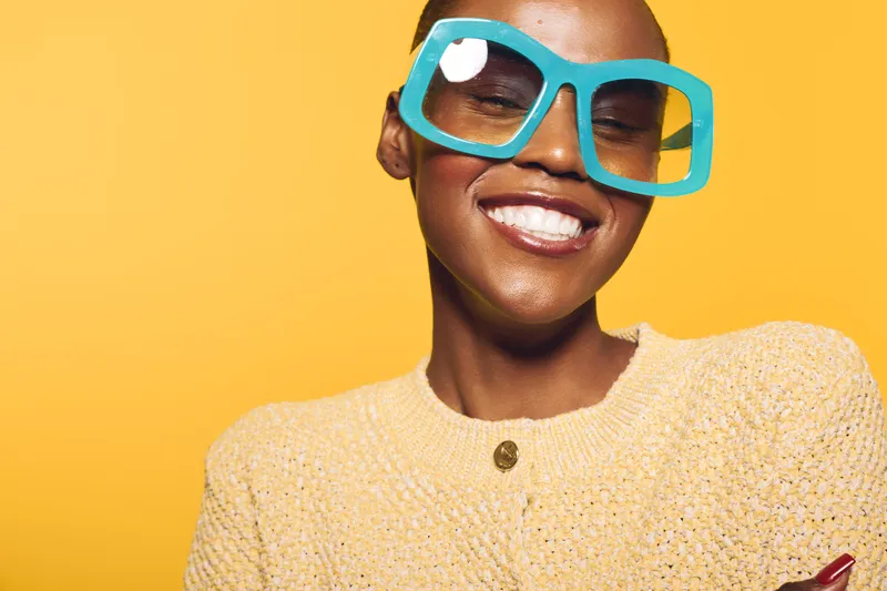 Smiling dark skinned African woman wearing large blue glasses and a yellow sweater poses against a bright yellow background. Concept of happiness, fashion, and vibrant style.