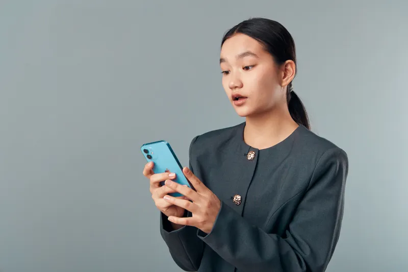 Young woman holding and looking at smartphone with surprised expression standing against plain background. Female in casual outfit using mobile phone and focused on device.