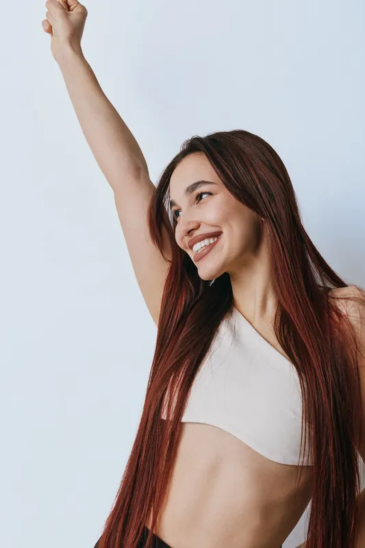 Young woman with long hair smiling and raising arm in celebration or victory against light background. Positive emotion, happiness and confidence in casual outfit and natural look.