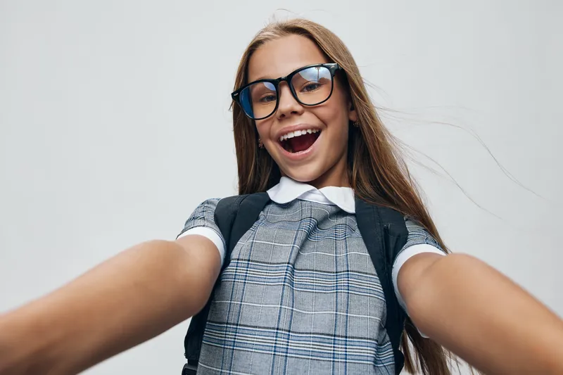 Girl child with glasses smiling happily in school uniform and backpack, posing for selfie with joyful facial expression and long hair on plain background.