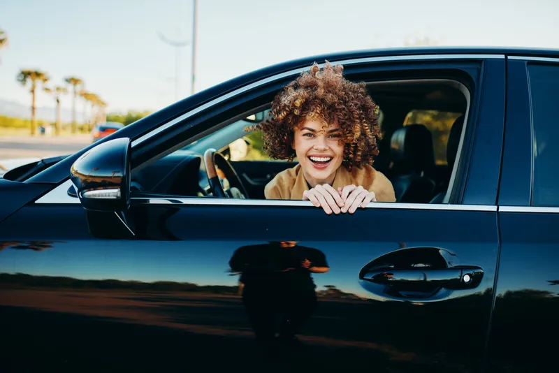 A young woman with curly hair smiles from the driver seat of a sleek dark car under a bright daytime sky.