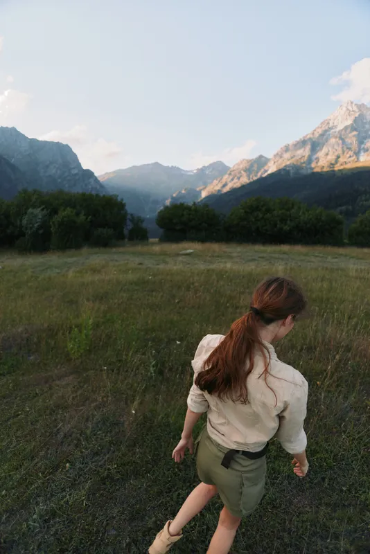 A woman with long hair walks through a green meadow surrounded by mountains under a clear sky. Lifestyle concept of nature exploration and outdoor activity.