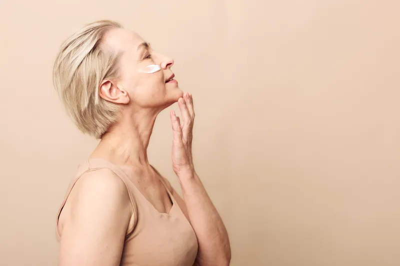 Profile of mature woman with short blonde hair applying face cream on cheek, eyes closed. Soft beige background and natural light create calm atmosphere. Concept of skincare and aging gracefully.