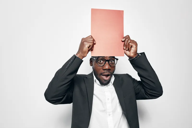 African American man with dark skin wearing glasses and black suit holding red folder above head with surprised expression on white background.