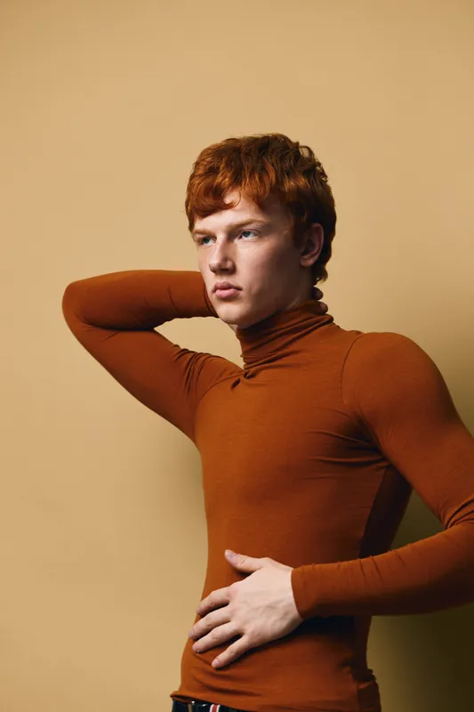 Young man with red hair wearing orange turtleneck posing with hand on neck and waist. Studio portrait with neutral background showing thoughtful expression and casual fashion style.