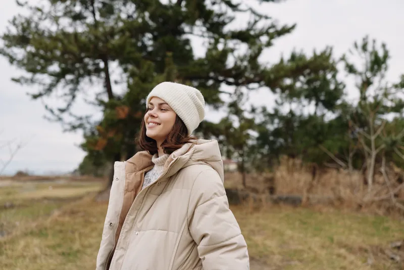 Woman in a warm beige coat and beanie smiles outdoors in a windy park, trees and open sky behind, capturing casual seasonal fashion and outdoor style.