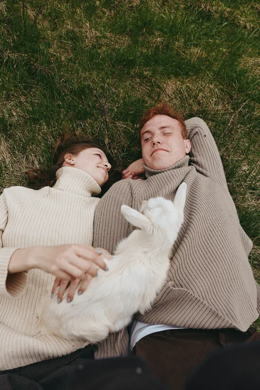 Two people lie on grass outdoors with a white goat kid, cozy sweaters, relaxed mood, gentle touch, intimate moment, in nature. Lifestyle scene highlighting relaxation, companionship, comfort, and