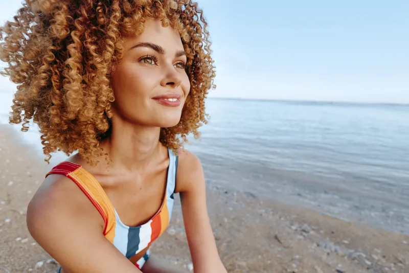 Young woman with curly blonde hair smiles thoughtfully while sitting on a sandy beach near the calm sea under a clear blue sky on a sunny day.